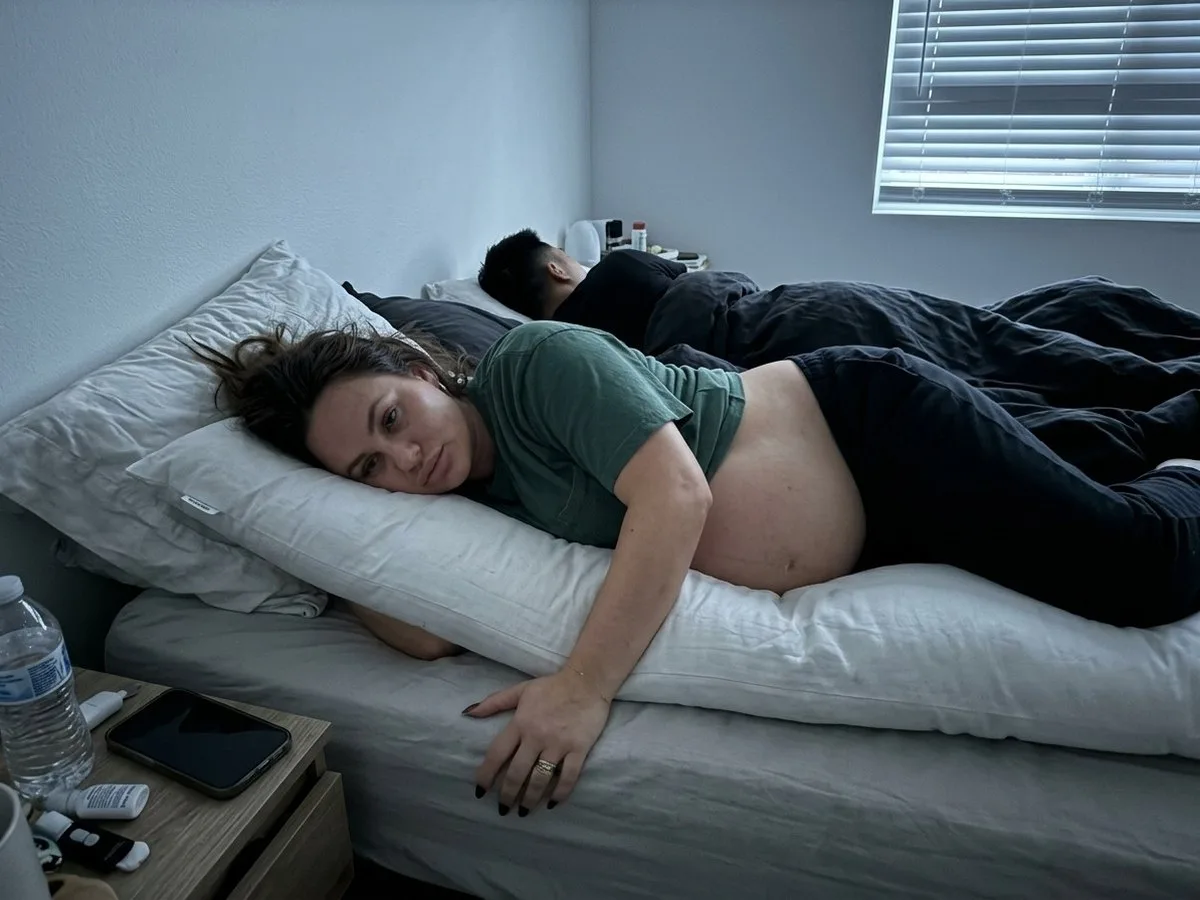 Pregnant woman awake before dawn sunk deep into a soft, flattened body pillow, her bump resting on the mattress
