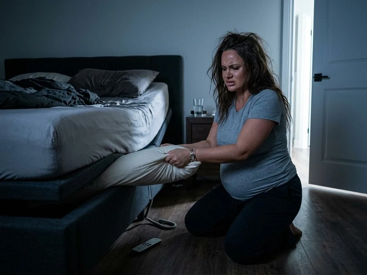 Pregnant woman kneeling at the side of an adjustable bed at night, pulling the end of a long pillow out of the gap between the mattress and the frame