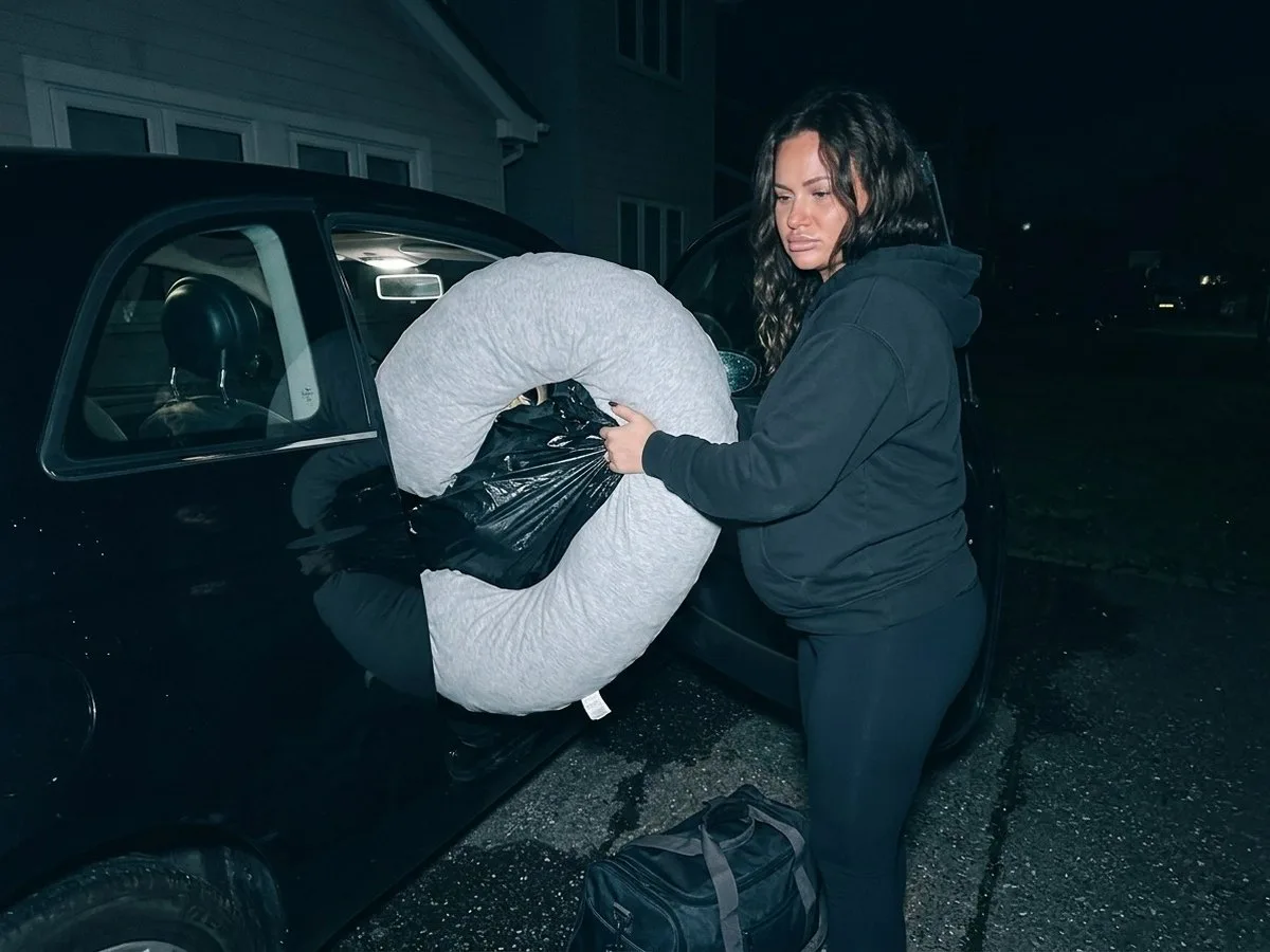 Tired pregnant woman at night in a driveway, struggling to push a large C-shaped pillow wrapped in a plastic bag into the back seat of a car