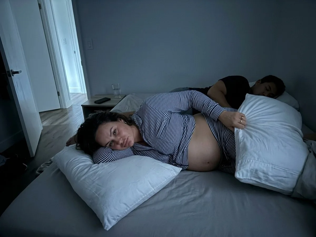 Pregnant woman awake on her side at night on a full bed, a tower of regular pillows collapsing behind her, partner asleep close beside her