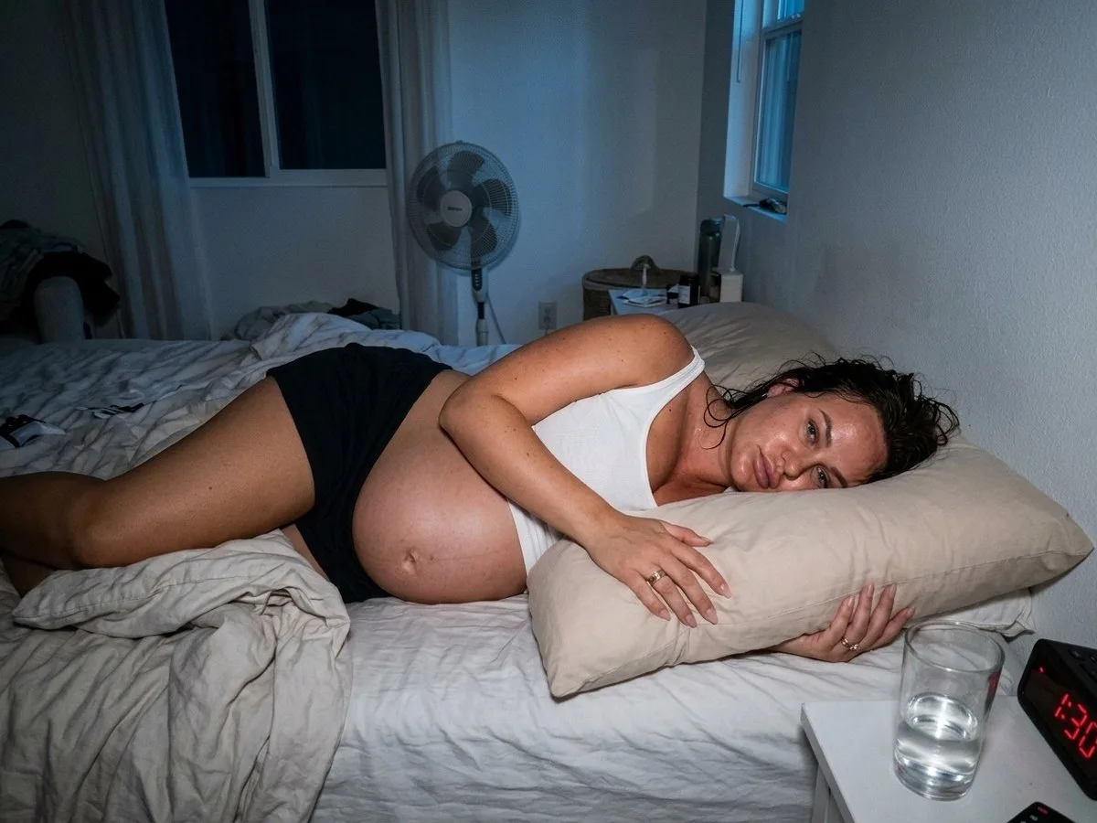 Pregnant woman awake on a hot summer night, peeling a damp pillow away from her bump with a fan running