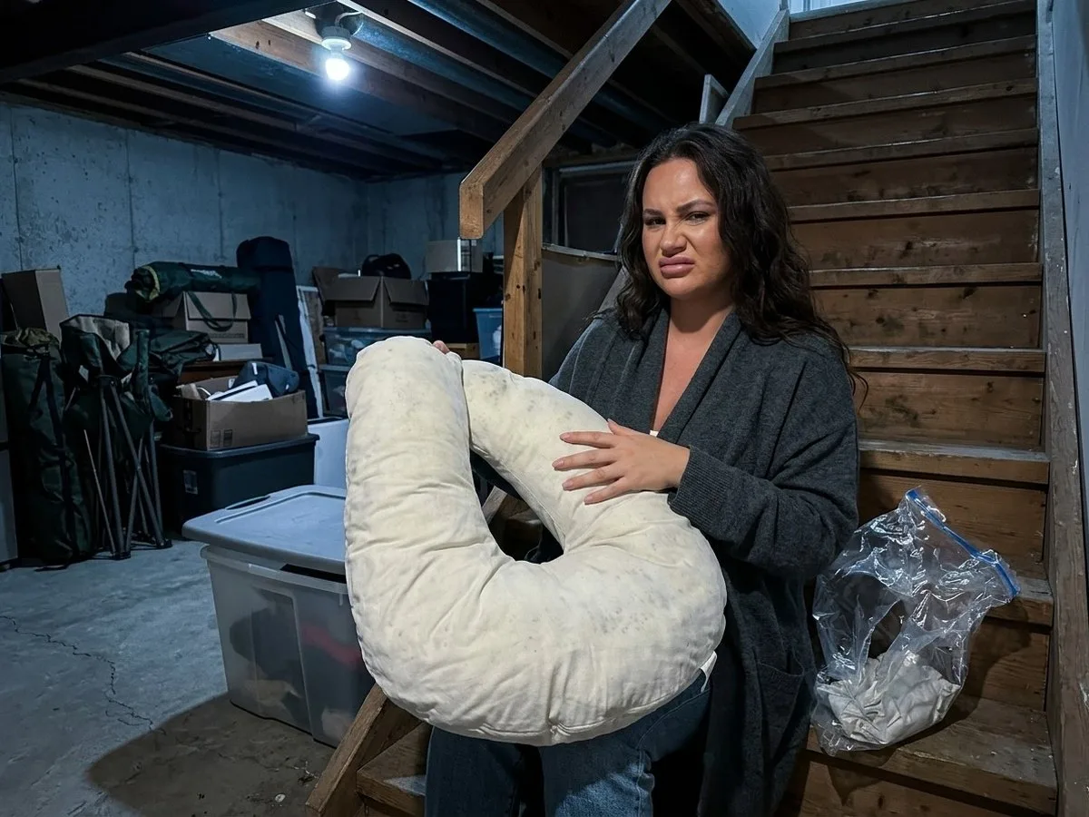 Pregnant woman sitting on basement stairs holding a flattened, musty pregnancy pillow just taken out of a cut-open vacuum bag