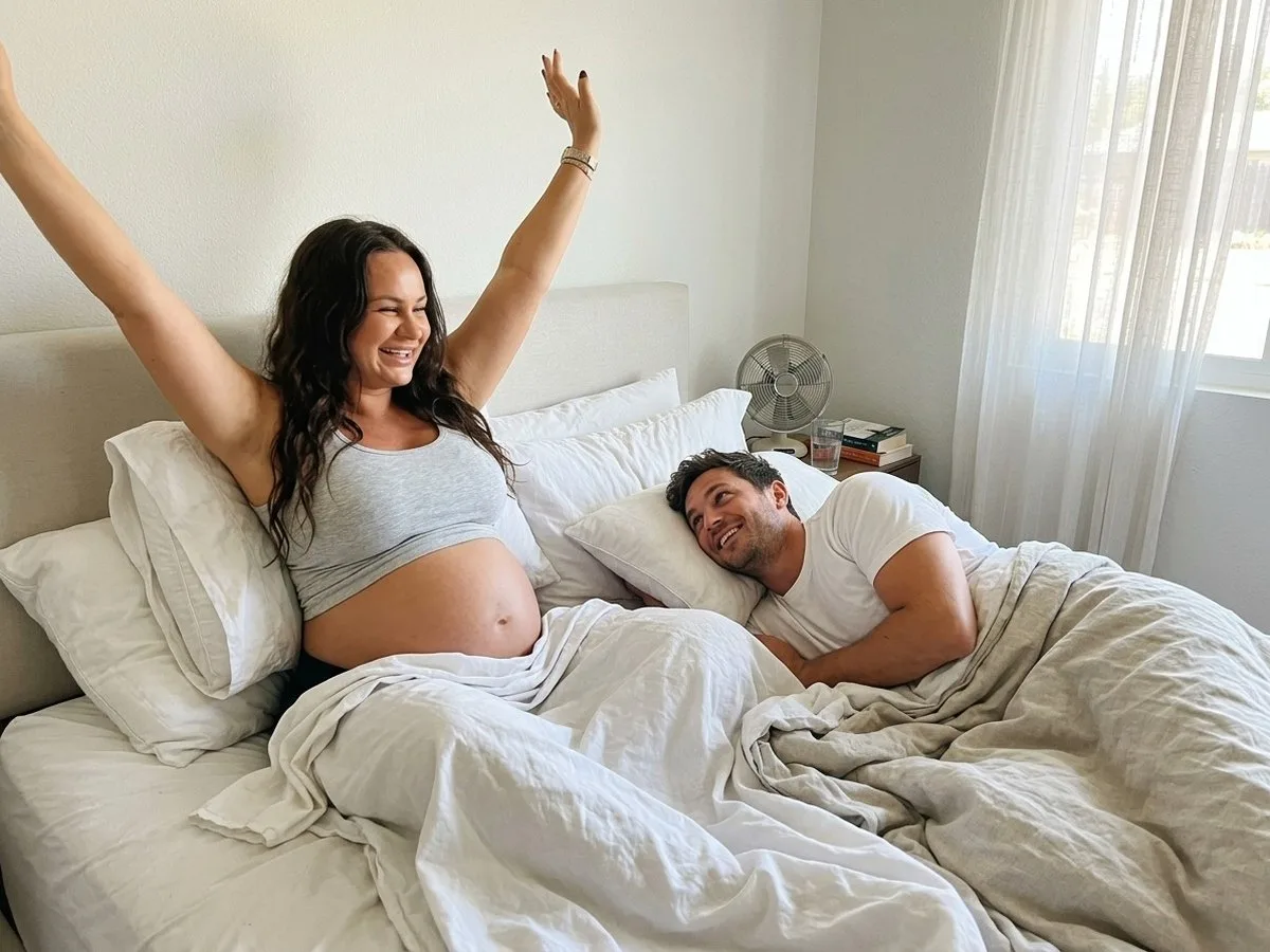 Bright summer morning, pregnant woman sitting up and stretching happily in bed under a light sheet while her partner, no longer in a hoodie, laughs beside her