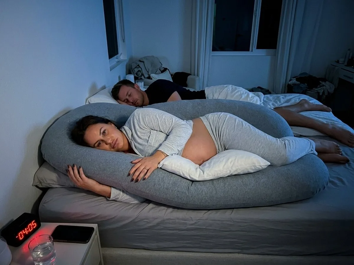 Pregnant woman awake at 4 a.m. inside a large C-shaped pillow with an extra pillow pushed under her bump, partner on the edge of the bed
