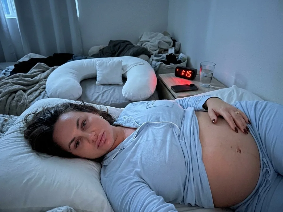 Pregnant woman lying on her right side at night, awake and tired, a one-sided body pillow left behind her on the other side