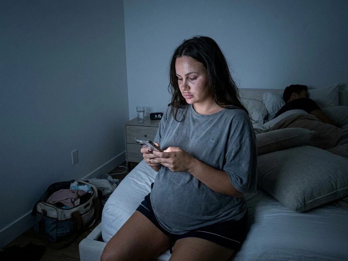 Heavily pregnant woman sitting on the edge of the bed at night, looking at a shopping cart on her phone, couch cushions piled behind her