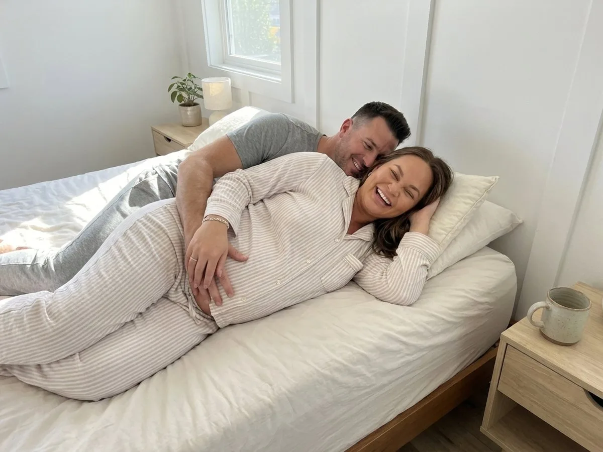 Sunny morning, pregnant woman laughing in bed while her partner hugs her from behind, both on a shared queen bed