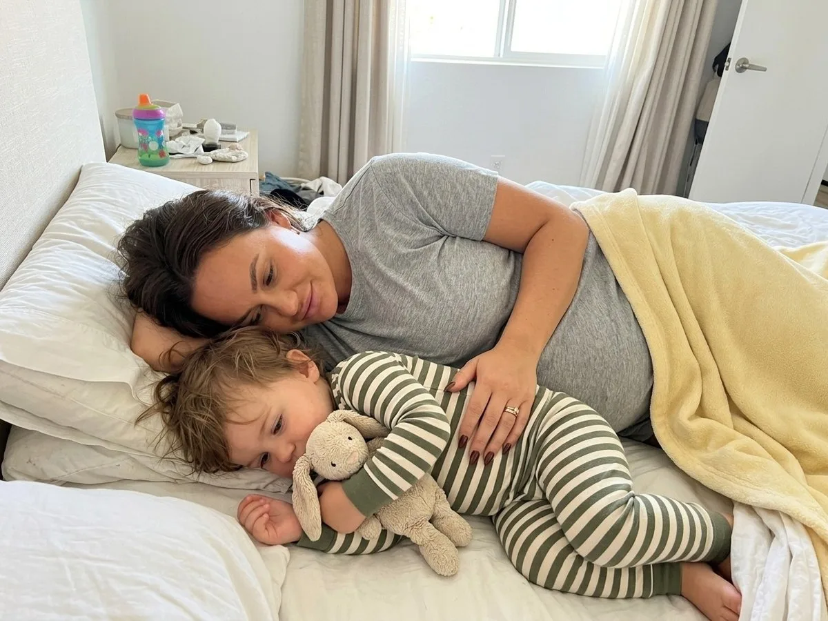 Saturday morning in bed, pregnant woman lying on her side smiling as her toddler curls up against her bump