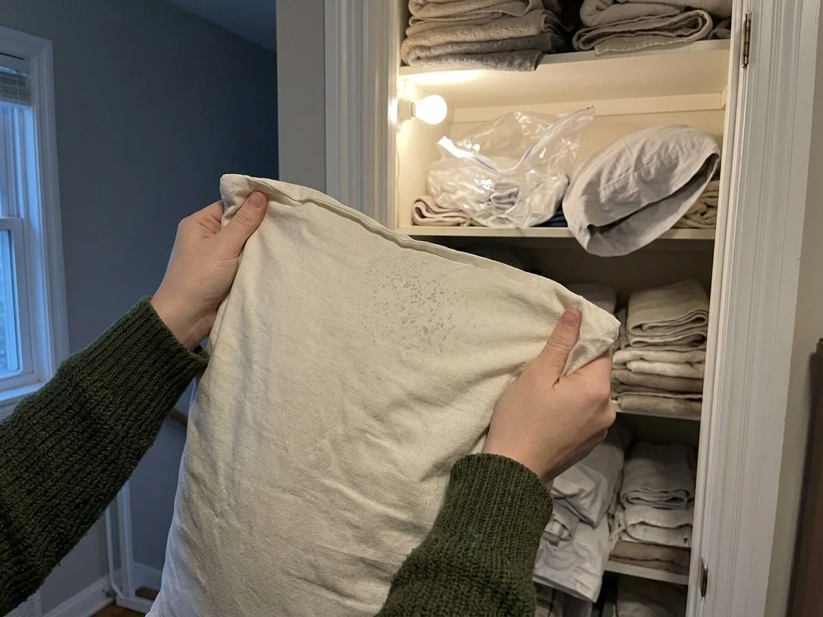 Pregnant woman sitting on a hallway floor in the evening with an unzipped storage bag and a pillow insert in her lap, looking at a patch of small grey spots