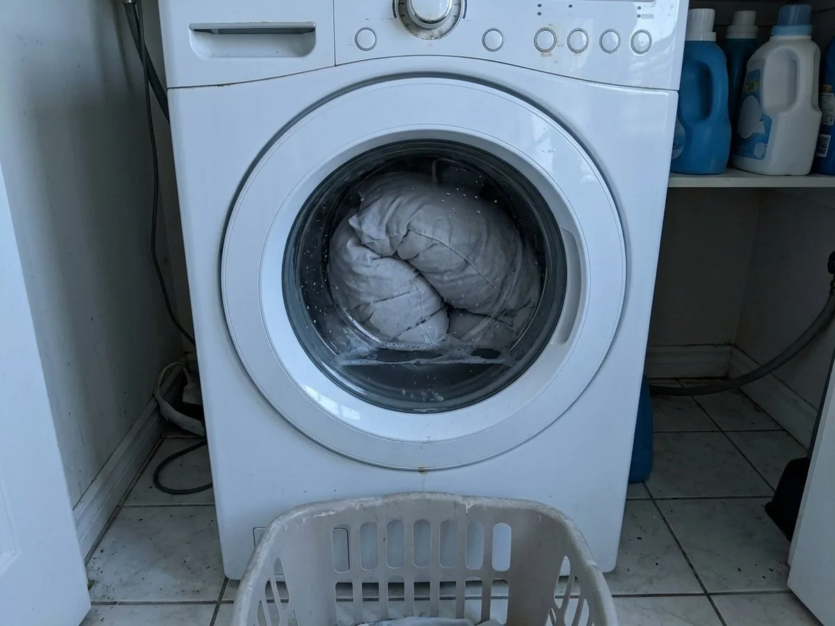 Pregnant woman crouching in front of a front-loading washing machine at night, looking at a blinking display, a swollen grey pillow pressed against the glass door