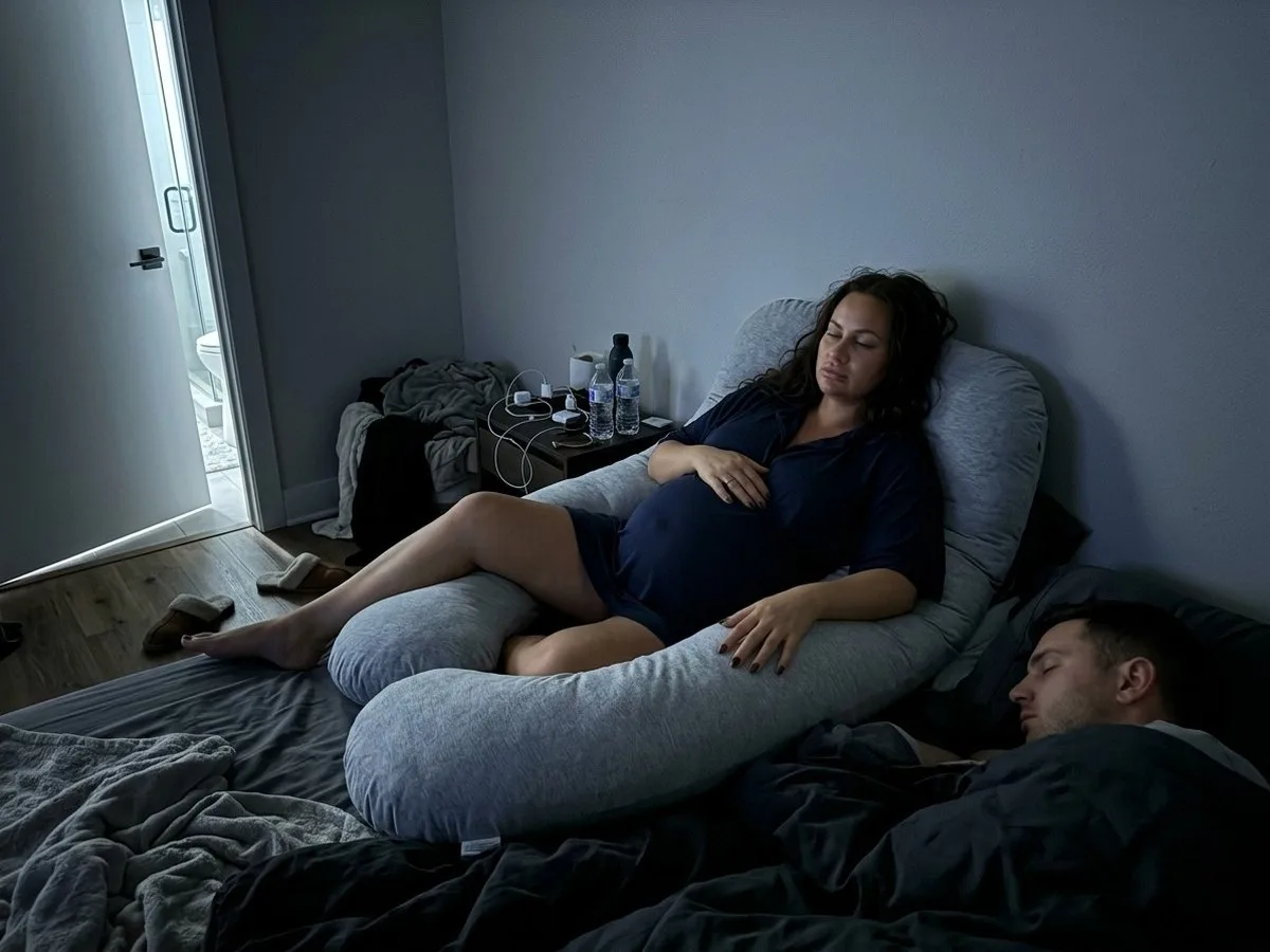 Pregnant woman at night sitting up inside a large U-shaped pillow on the wall side of the bed, one leg lifted over the pillow, her partner asleep on the outer side