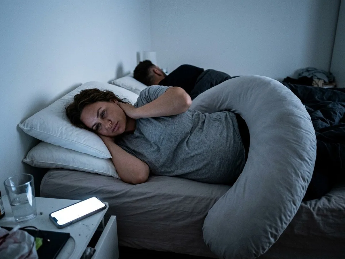 Tired pregnant woman awake in bed at night, a large pregnancy pillow pushed half off the bed and ordinary pillows stacked under her head
