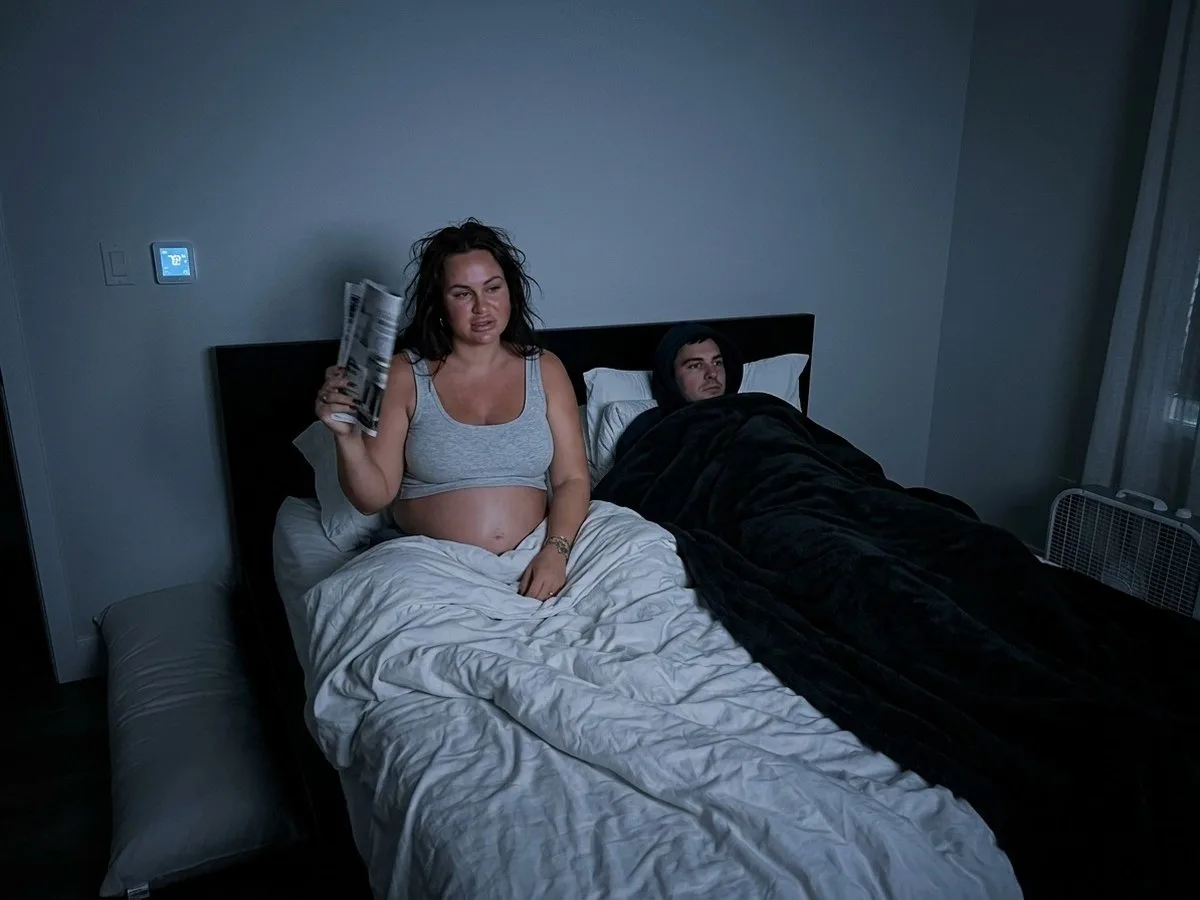 Night, pregnant woman sitting up in bed fanning herself while her partner shivers in a hoodie under a thick blanket
