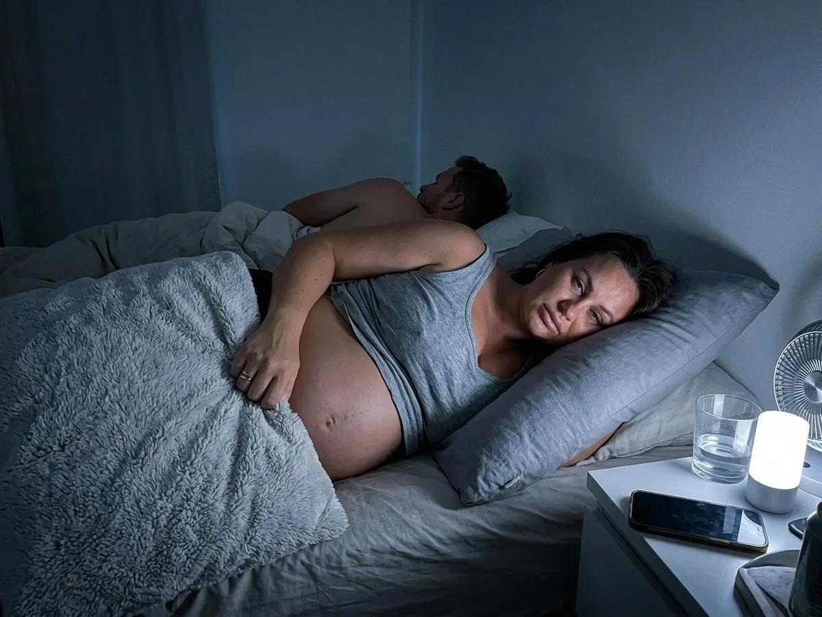 Pregnant woman awake at night pulling a thick plush pillow cover away from her belly, a fan on the nightstand