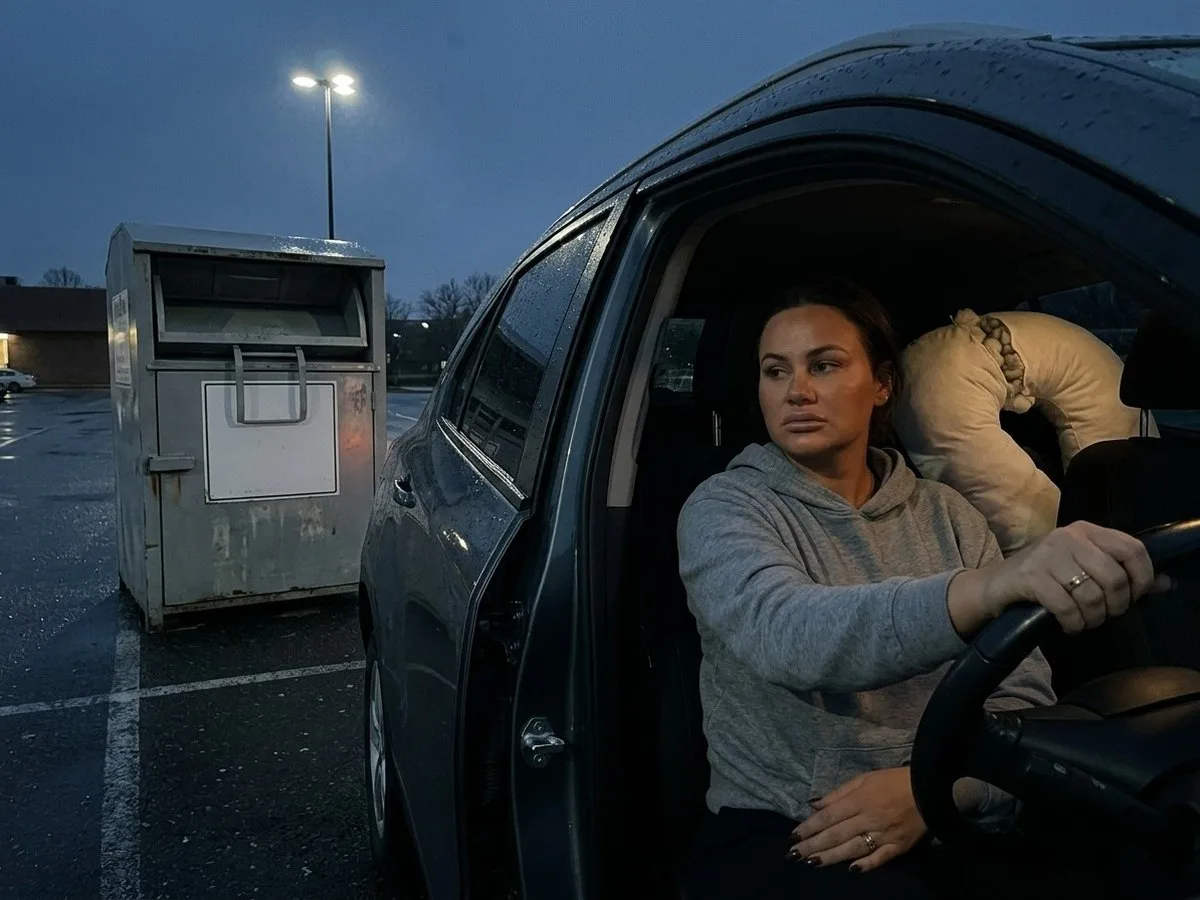 Woman sitting in a parked car at dusk beside a textile donation bin, a large worn pregnancy pillow with a split seam on the back seat