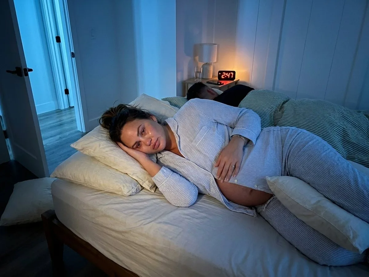Exhausted pregnant woman awake on her side at night, pillows scattered and one on the floor, bedside clock glowing 2:47