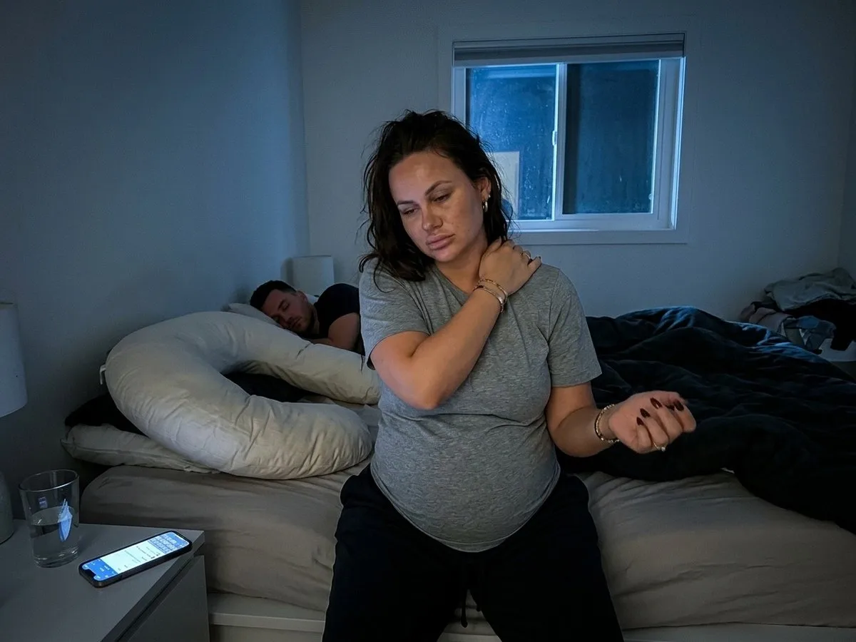 Pregnant woman sitting on the edge of the bed at night shaking out a numb hand, a C-shaped pillow behind her