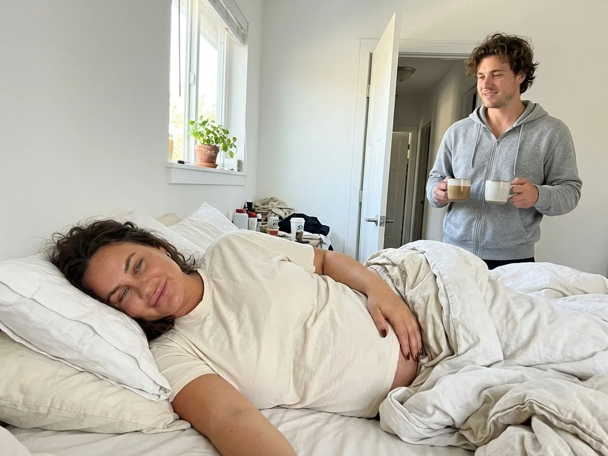 Bright Saturday morning, pregnant woman asleep and smiling on her side while her partner stands in the doorway holding two coffee mugs