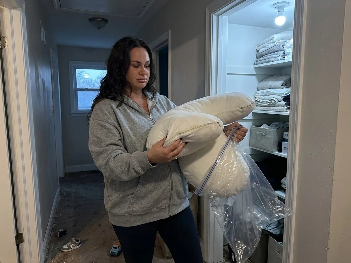 Pregnant woman in a dim hallway at night holding an old pregnancy pillow just taken out of a storage bag, looking unsure