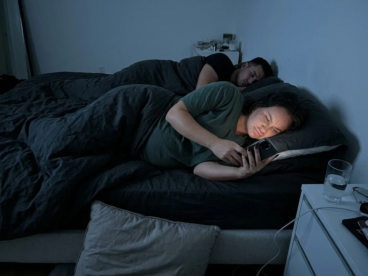 Pregnant woman lying on her side in a dark bedroom late at night, squinting at pillow listings on her phone while her partner sleeps
