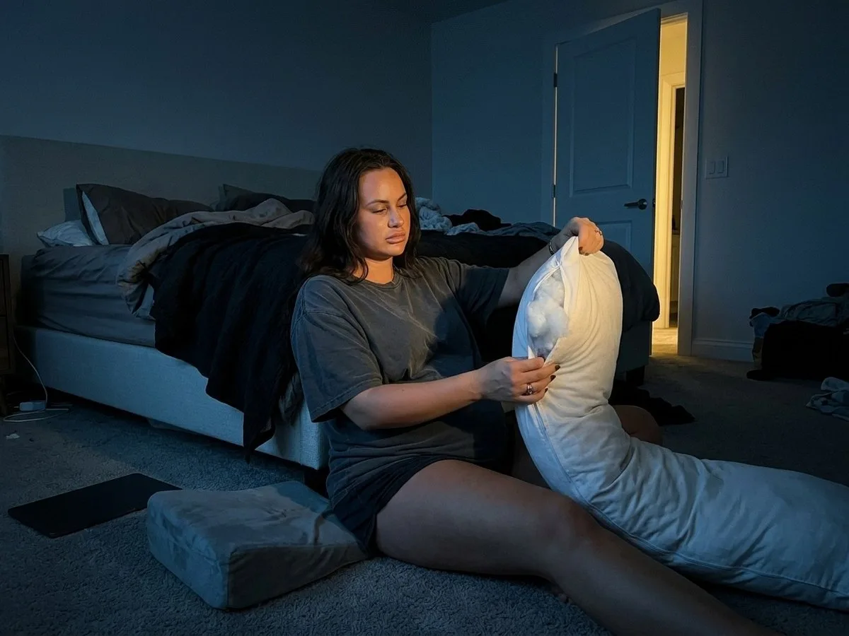 Pregnant woman at night sitting on the bedroom floor next to two cheap pregnancy pillows, holding an open seam with filling showing