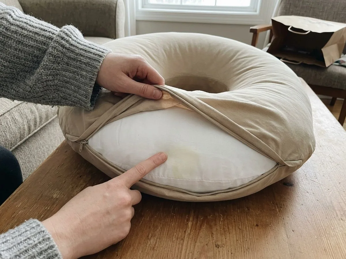 Pregnant woman sitting on a living room floor with an unzipped pillow cover pulled back, inspecting a stain on the inner pillow