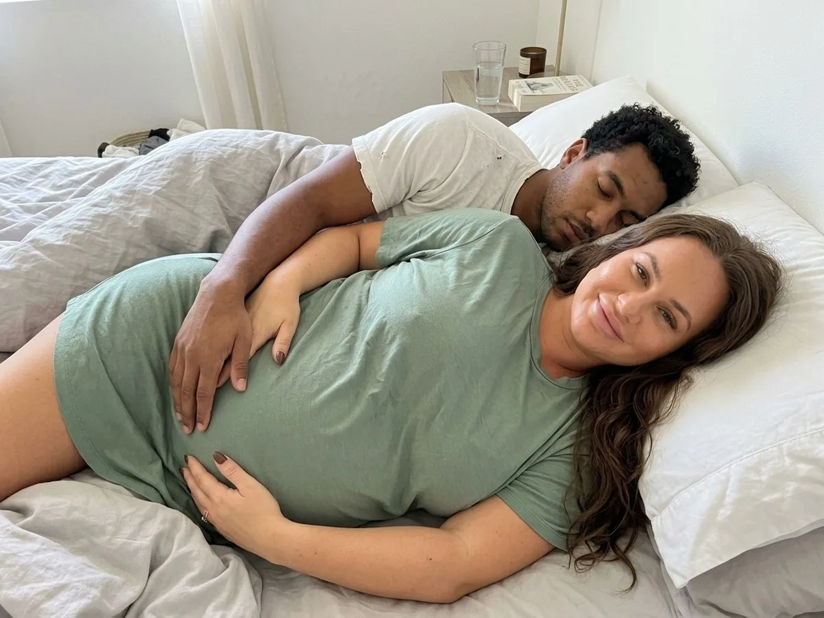 Sunny morning in a full-size bed, pregnant woman awake and smiling while her partner sleeps close with his arm across her shoulder