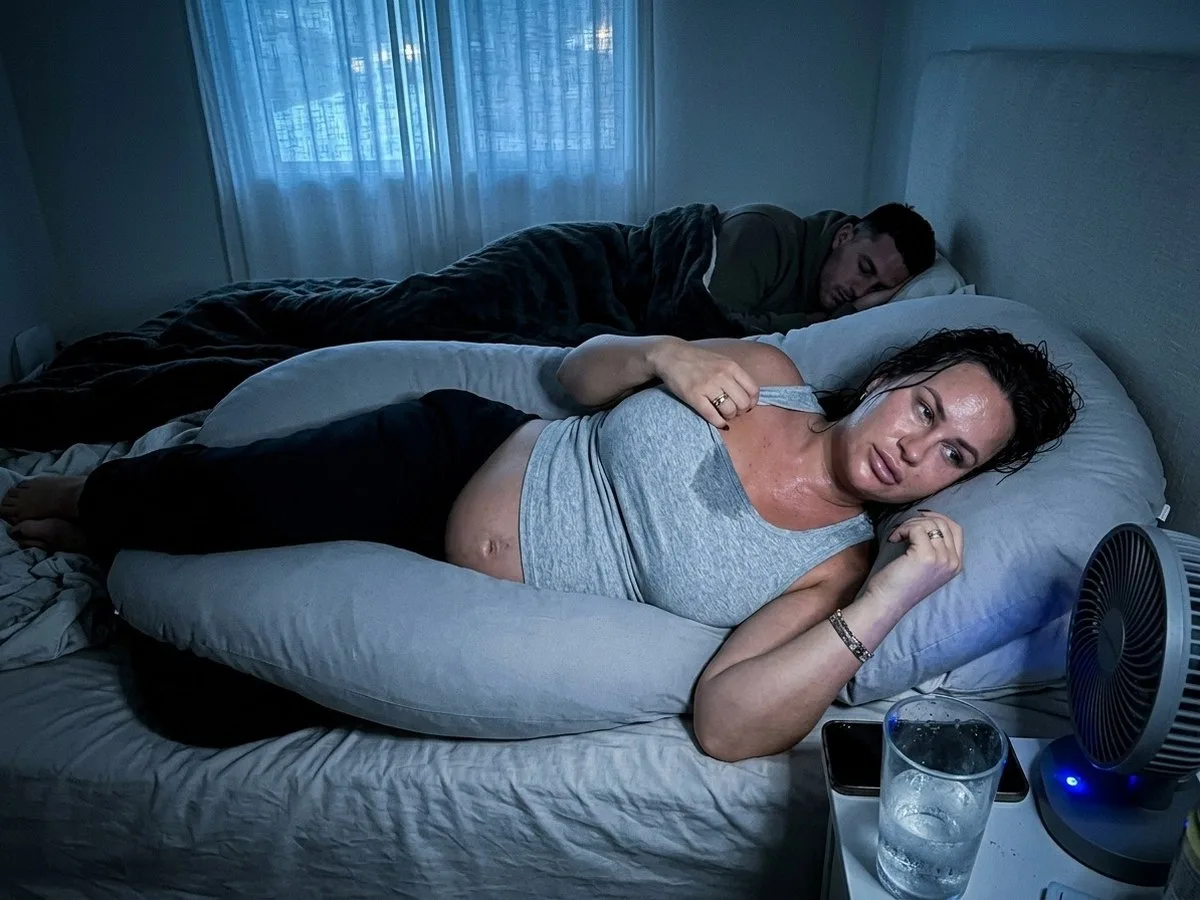 Hot summer night, pregnant woman awake and sweating inside a large U-shaped pillow, sheet kicked off, a fan blowing at her