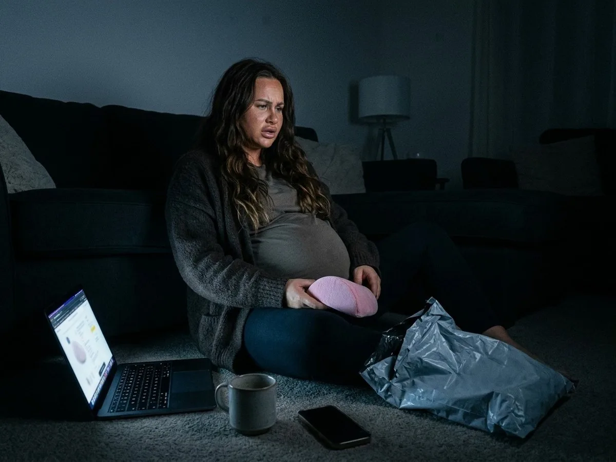 Pregnant woman sitting on the floor at night holding a single pink wedge cushion next to an opened shipping bag, looking frustrated at her laptop