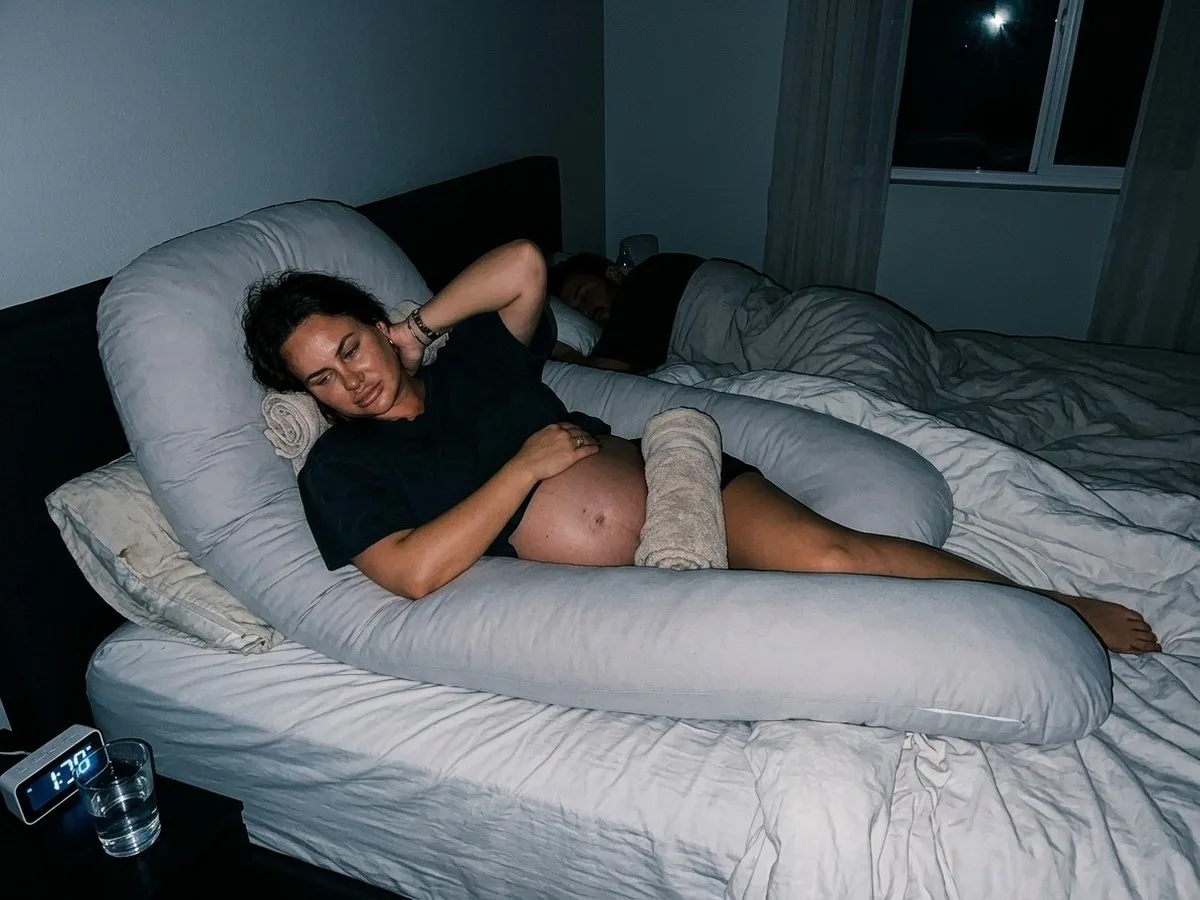 Night, a petite pregnant woman lying in a long U-shaped pillow that is too big for her, her head pushed up by the top curve and rolled towels stuffed around her