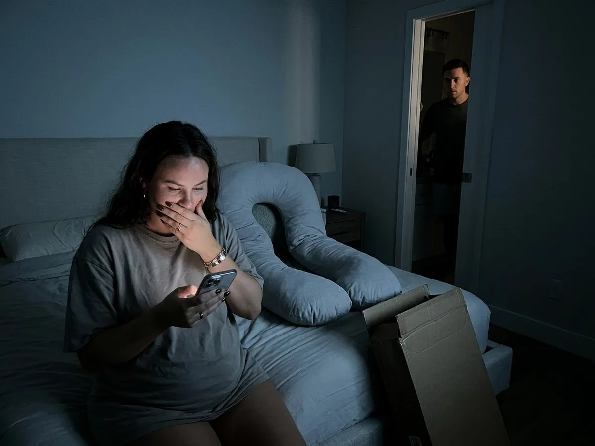 Pregnant woman sitting on the edge of the bed at night reading a long returns page on her phone, an oversized pillow slumped behind her