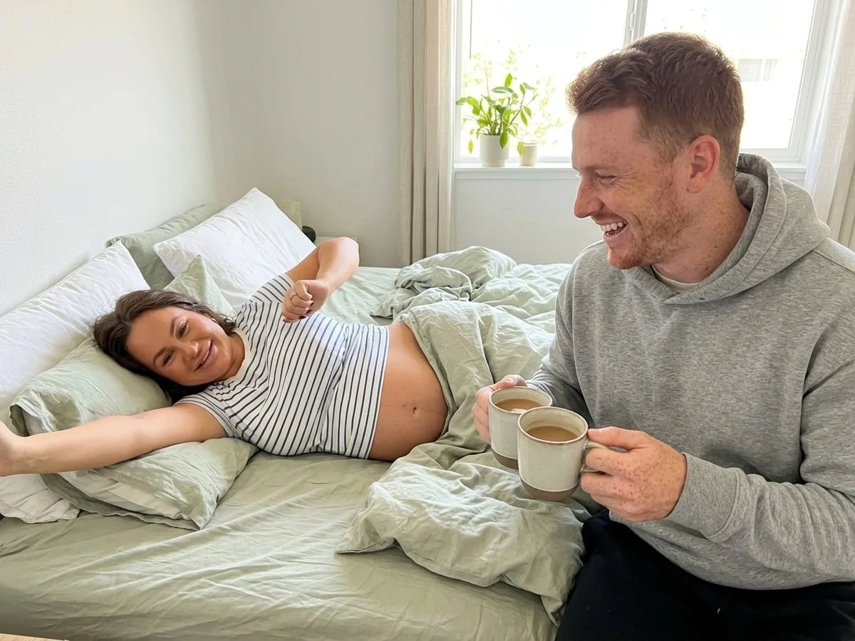 Sunny Sunday morning, pregnant woman waking up smiling as her husband sits on the edge of the bed holding two mugs of coffee