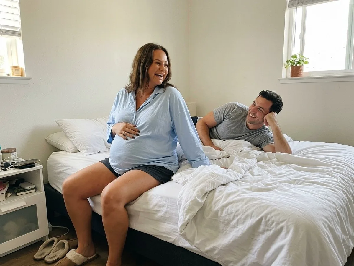 Sunny morning, pregnant woman laughing as she sits up on the edge of the bed with her feet on the floor while her partner smiles from his side of the same bed