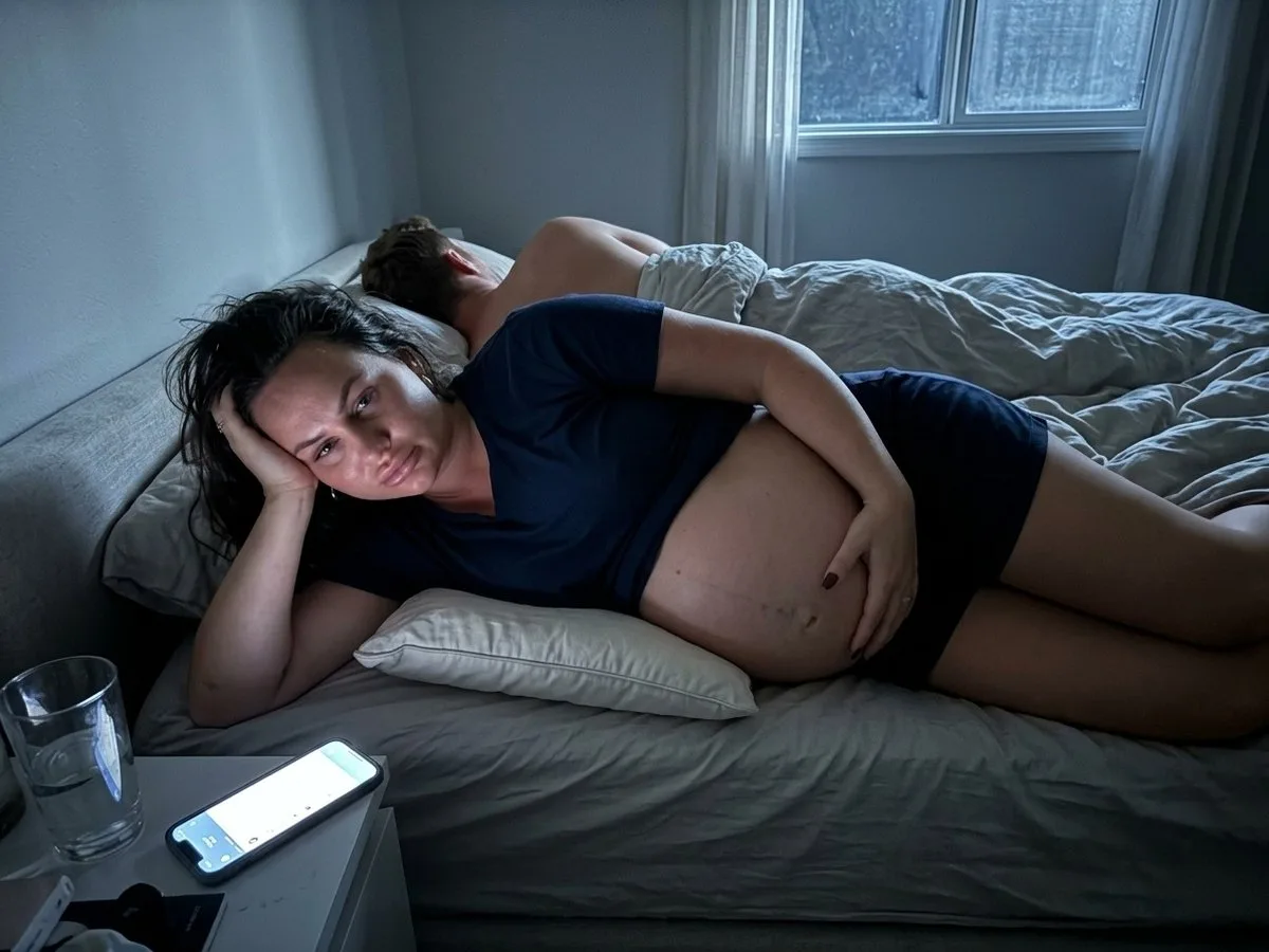 Pregnant woman with a large twin bump awake on her side at night, holding her belly where it hangs off a flattened pillow