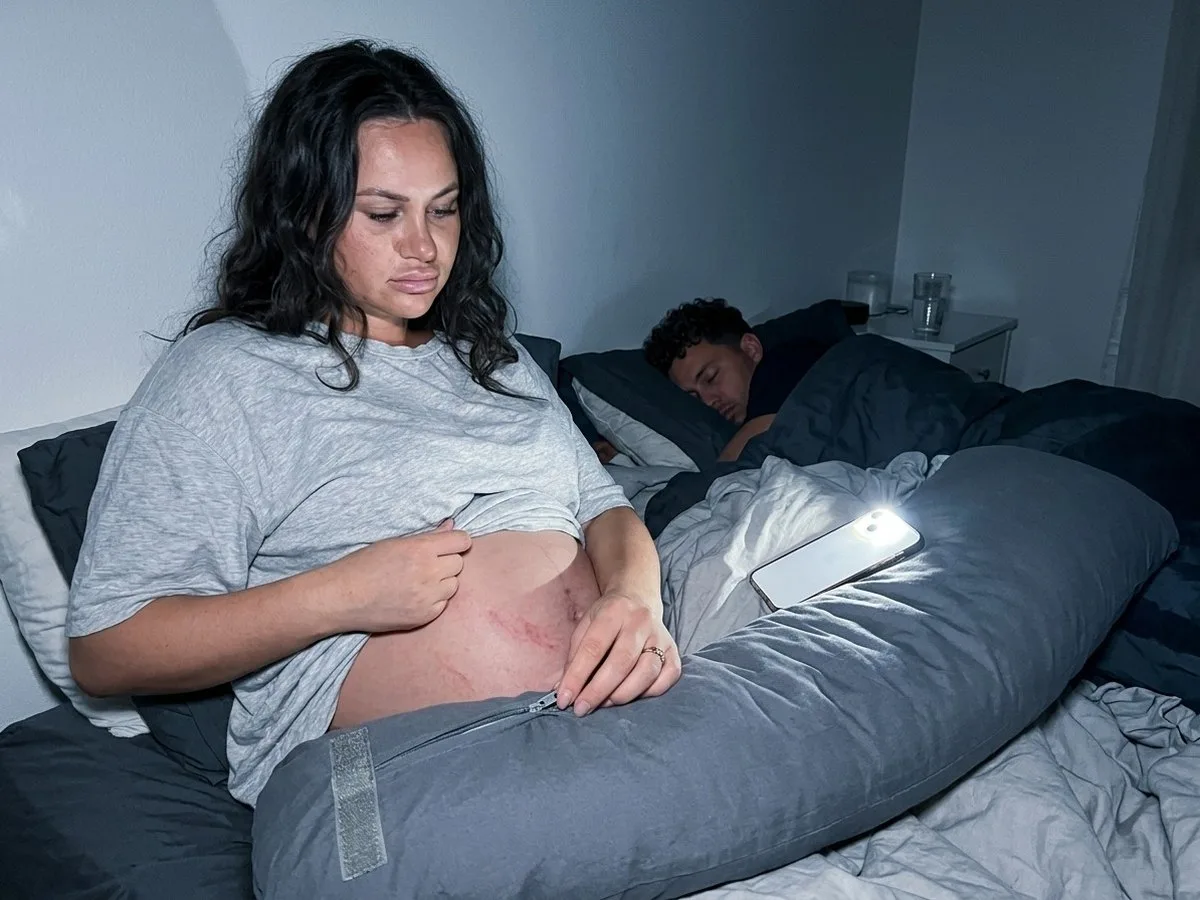 Pregnant woman awake at night, rubbing a red mark on her side and frowning at the zipper on her pregnancy pillow