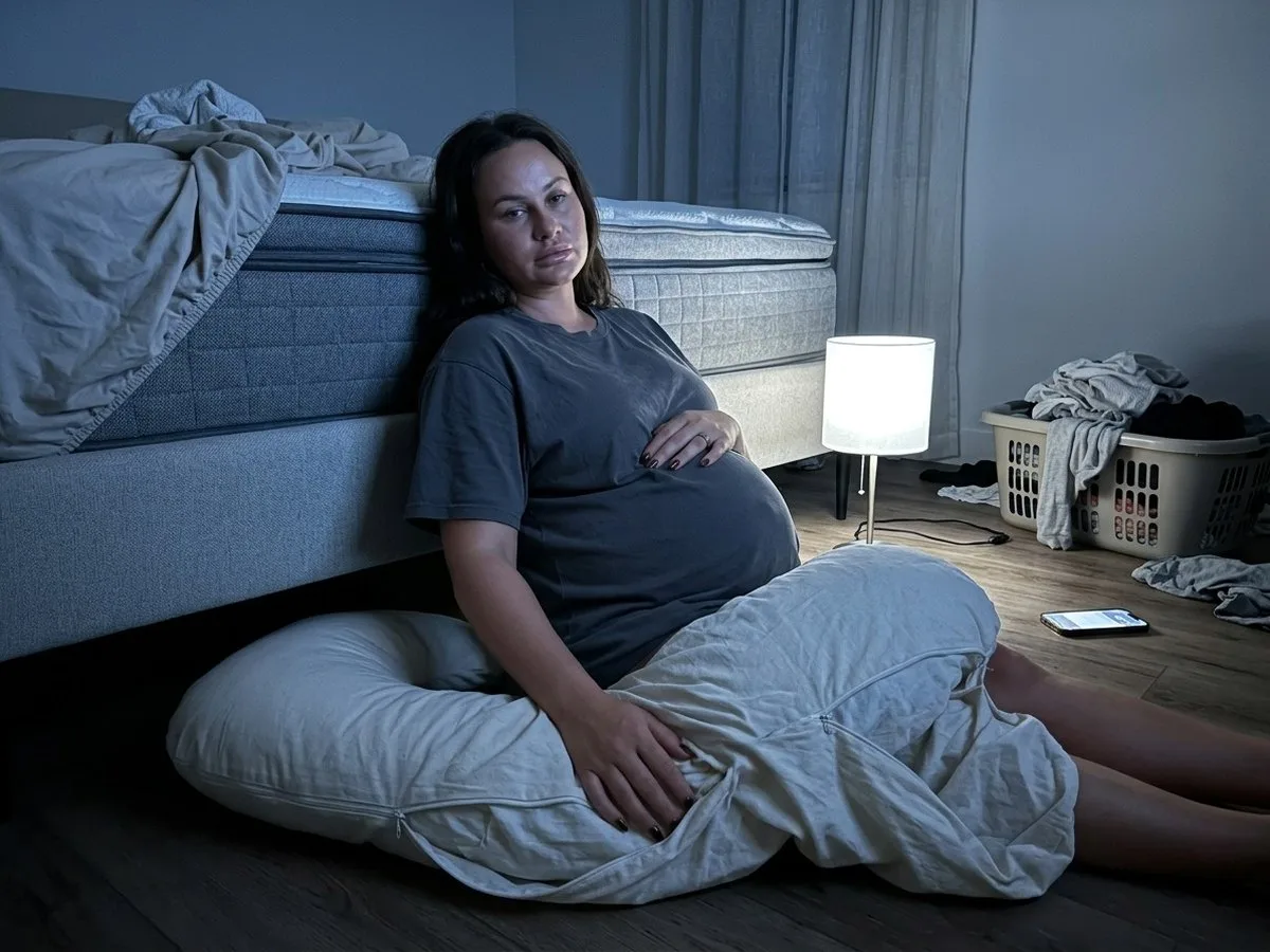 Pregnant woman sitting on the bedroom floor at night beside a long U pillow she has pulled off the bed, looking up at the stripped mattress