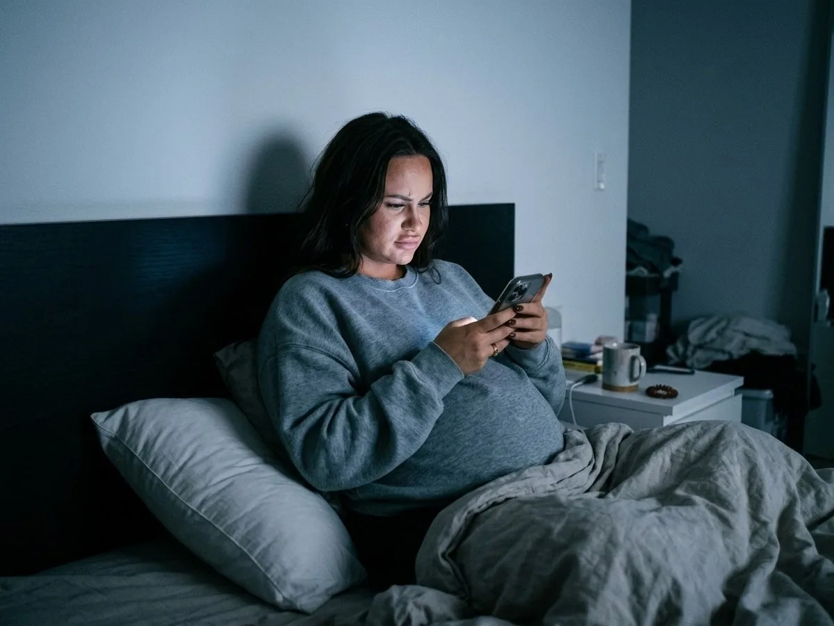 Pregnant woman sitting up in bed at night replaying a video on her phone, frowning, a plain long body pillow lying beside her