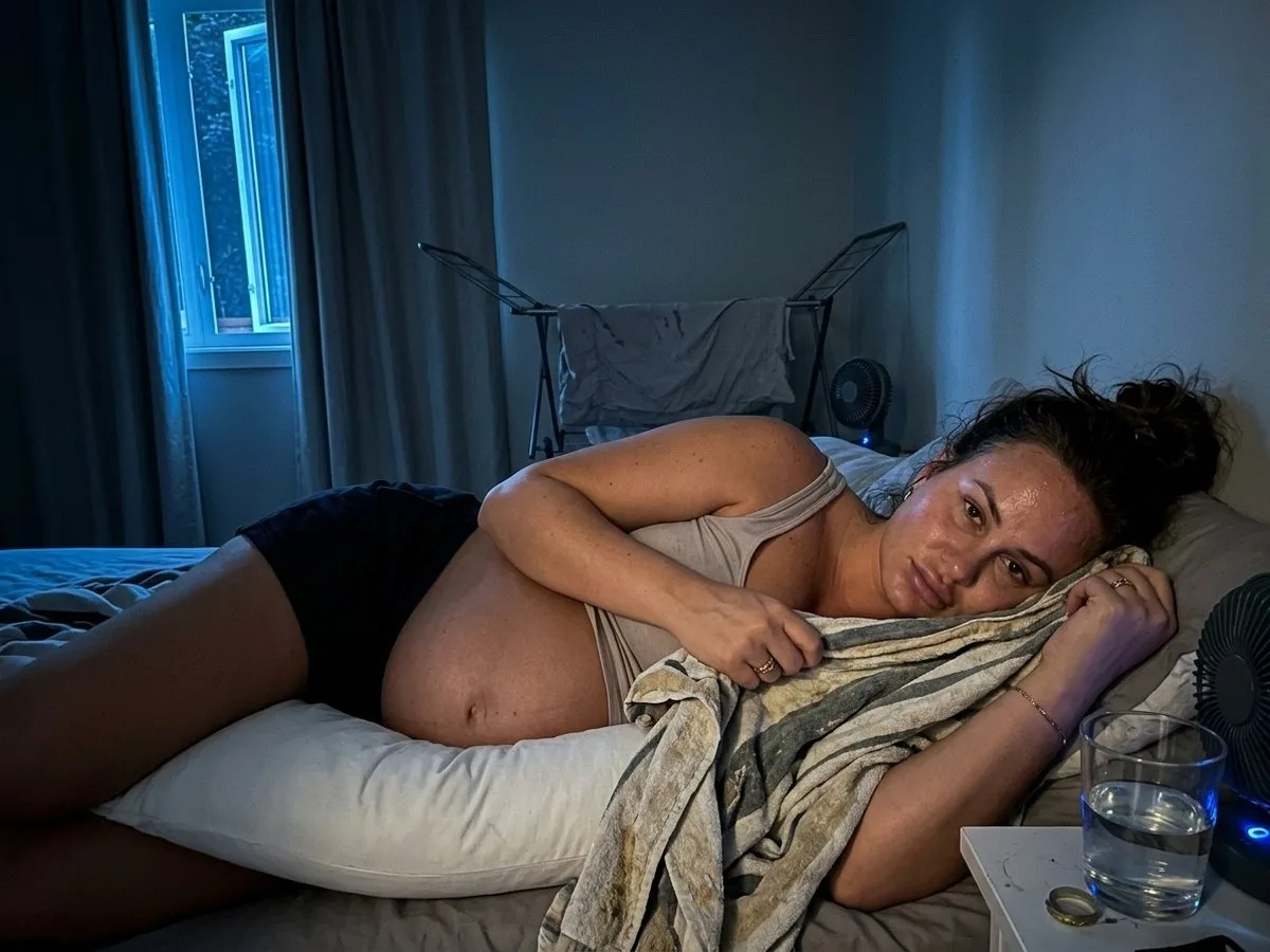 Pregnant woman at night on a summer bed, lying on a bare pillow insert with a beach towel slipping off it, a damp cover on a drying rack behind her