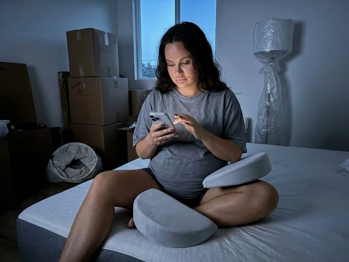 Pregnant woman sitting on a mattress among moving boxes at night, holding two loose wedge cushions with no strap, looking at a product photo on her phone